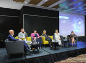 Sześć osób, cztery kobiety i dwóch mężczyzn, siedzi na scenie w fotelach podczas dyskusji panelowej. Duży ekran za nimi wyświetla tekst i grafikę w języku polskim. Pomiędzy krzesłami umieszczono małe stoliki z butelkami wody i szklankami. Otoczenie jest nowoczesne i profesjonalne.