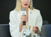 Kobieta o prostych blond włosach, ubrana w biały garnitur, siedzi na szarym krześle i trzyma mikrofon. Podczas mówienia gestykuluje jedną ręką. Na stole przed nią znajduje się przezroczysta karafka z wodą i szklanka. Tło jest ciemne i proste.