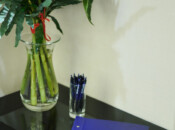 Szklany wazon ze słonecznikami i zielenią stoi na czarnym stoliku obok przezroczystego kubka z niebieskimi długopisami. Z przodu leży niebieska księga gości z napisem Księga Pamiątkowa z białą ilustracją i niebieskim godłem na okładce.