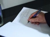 Osoba pisze długopisem na kartce białego papieru, którą trzyma drugą ręką. Tekst jest częściowo nieczytelny. Scena rozgrywa się na stole z ciemnego szkła, a w tle widoczna jest szklanka z wodą i uchwyt na długopis.