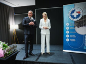 Mężczyzna w ciemnym garniturze przemawia do mikrofonu na scenie obok kobiety w białym garniturze. Stoją obok niebieskiego baneru o tematyce medycznej i stołu z bukietami kwiatów i trofeami. Sceneria wydaje się być formalnym wydarzeniem lub ceremonią w pomieszczeniu.