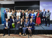Duża grupa profesjonalnie ubranych mężczyzn i kobiet pozuje razem na scenie w sali konferencyjnej. Niektóre osoby siedzą z przodu, a inne stoją za nimi. W tle widoczny jest ekran prezentacyjny i baner.